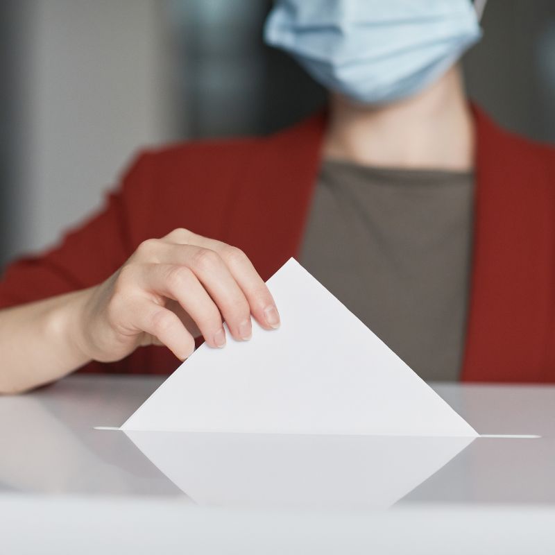 personne masquée qui met un bulletin de vote dans une urne